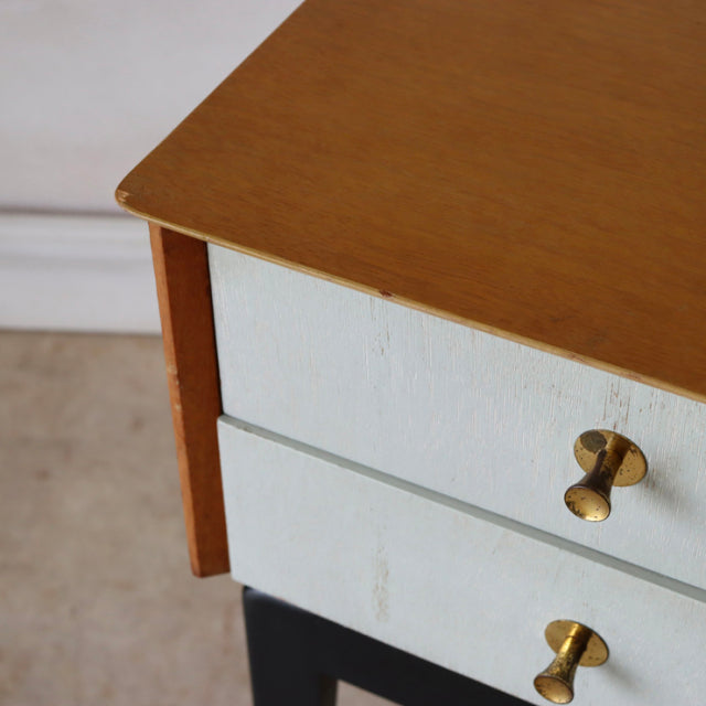Mid-century British teak bedside cabinet with light blue drawers, brass knob handles, and black legs featuring visible wood.