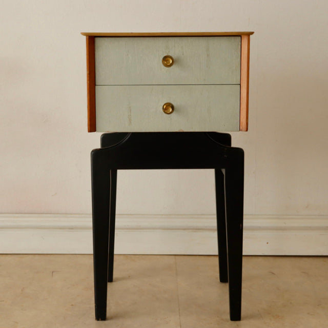 Vintage British mid-century teak bedside cabinet with two painted drawers and brass knobs on black tapered legs.