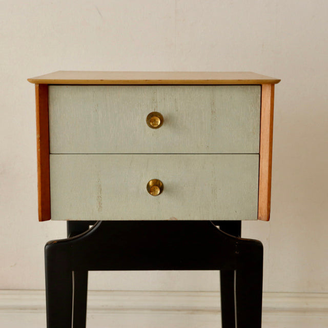 Mid-century British vintage teak bedside cabinet with light green drawer fronts, brass knobs, and slender black legs.