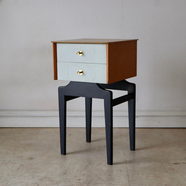Mid-century British vintage teak bedside cabinet with light drawer fronts and brass handles on black wooden stand.