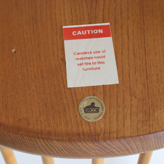 Close-up of vintage ERCOL Quaker chair seat in natural teak, showing wood grain and branded circular metal badge embedded.