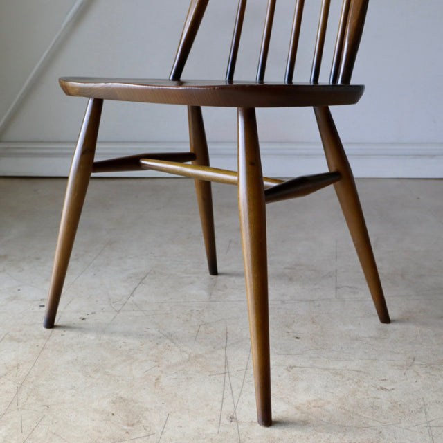 Mid-century British ERCOL Quaker chair in teak with slender spindle back, tapered legs, and visible wood grain in warm brown .