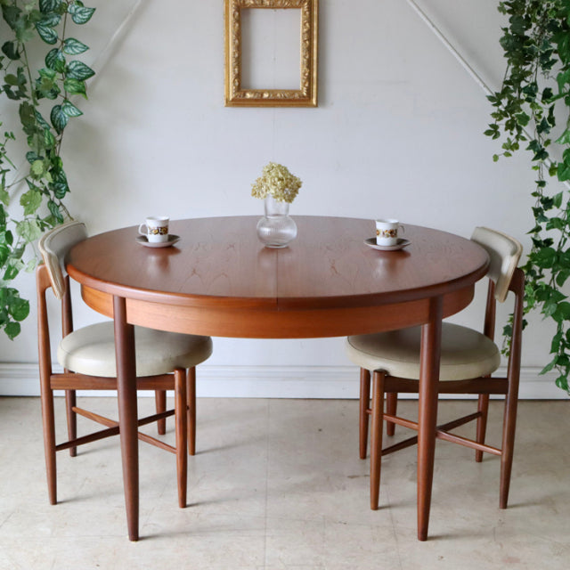 Vintage teak G-Plan mid-century British dining table with rounded top and tapered legs, paired with beige cushioned chairs.