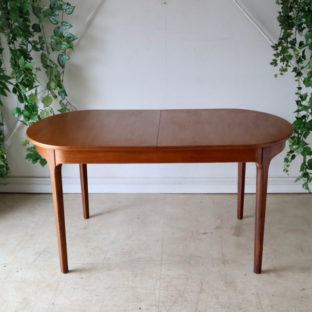 Mid-century British vintage teak dining table with oval top, slender tapered legs, rich wood grain finish.