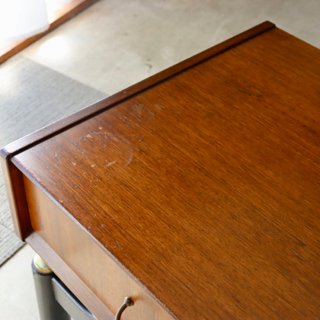 Vintage British mid-century teak bedside cabinet with warm wood grain and brass pull handle on slender legs.