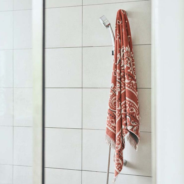 Red bandana-patterned cotton pile blanket with fringe edges hanging on a showerhead against white tile wall.
