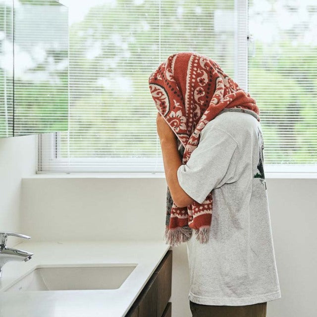 Person drying face with red vintage bandana-patterned cotton pile blanket in bright, minimalist bathroom setting.