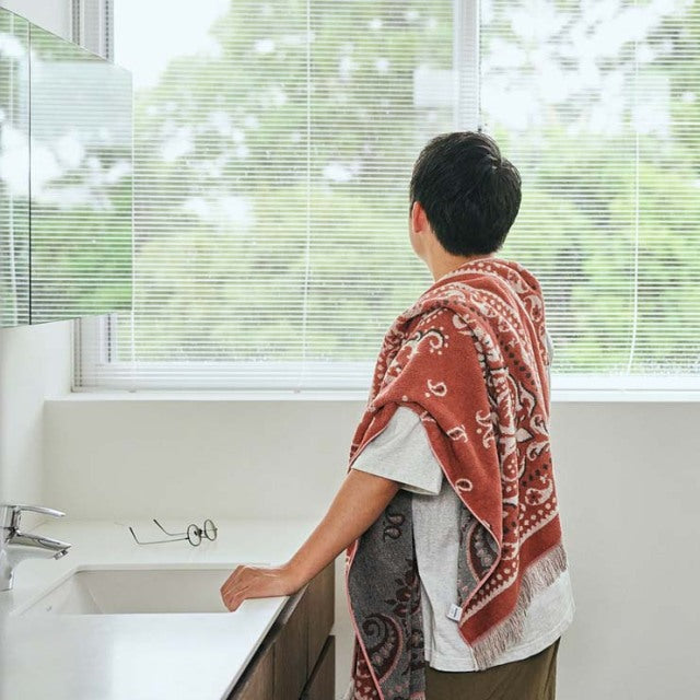 Man draped in a red bandana-patterned cotton pile blanket, standing by a white sink with a large window showing greenery in.