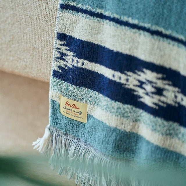Blue vintage-style cotton pile blanket with Native Border pattern and fringed edges draped over beige mid-century teak.