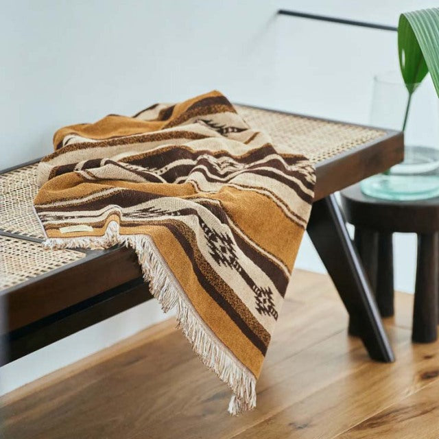 Mid-century teak bench with woven cane seat draped with a brown Native Border cotton pile blanket featuring fringe edges,.