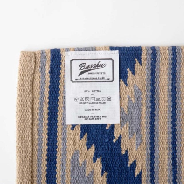 Close-up of a cotton rug with beige and navy blue zigzag stripes, textured weave, and a Basshu brand label sewn on,.