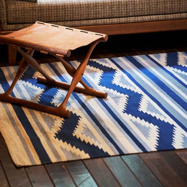 Mid-century teak folding stool with leather straps on vintage British-style cotton rug featuring blue and beige zigzag.