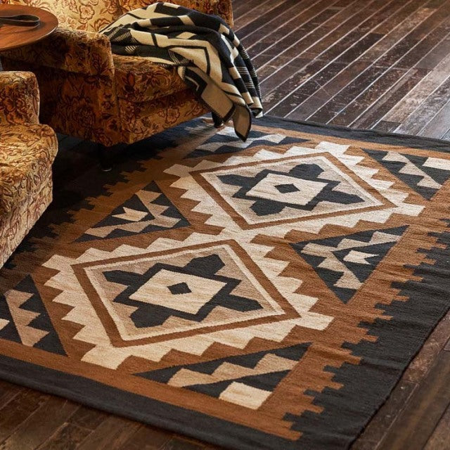Vintage British mid-century cotton rug with geometric patterns in brown, cream, and black on dark wooden floor beside teak.