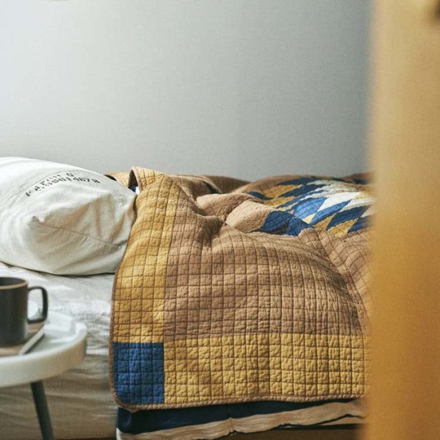 Camel patchwork quilt with vintage British mid-century design, featuring textured stitching and muted blue accents on a bed.