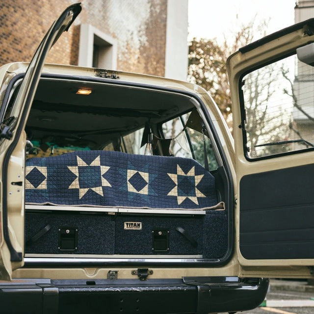 Blue patchwork quilt with vintage British design draped over teak mid-century storage trunk inside open classic vehicle.