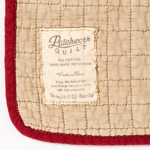 Close-up of beige cotton patchwork quilt with red border and vintage-style sewn label reading "Patchwork Quilt All Cotton.