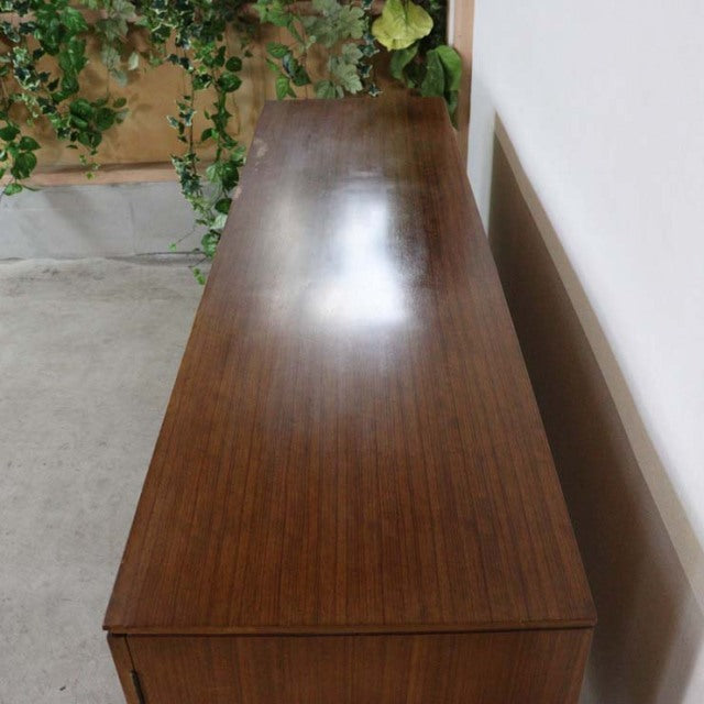 Long vintage British mid-century teak sideboard with smooth wood grain surface and tapered legs against a white wall.