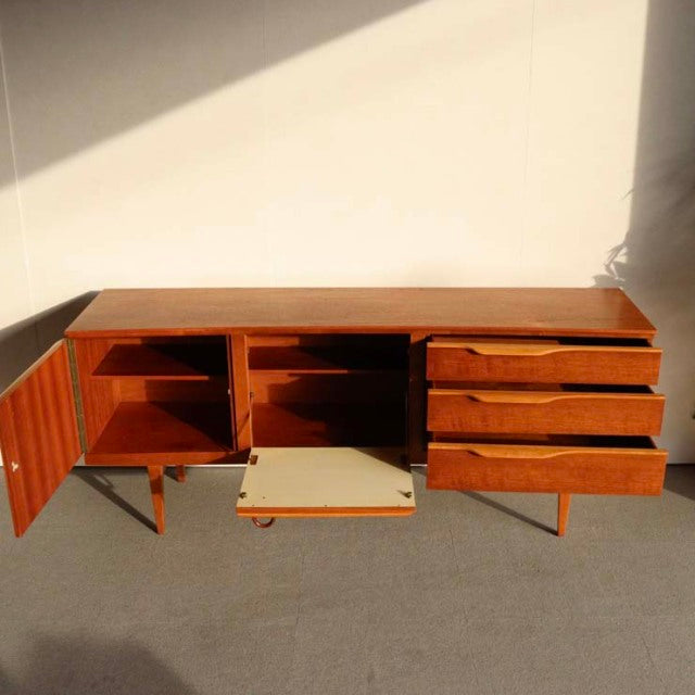 Mid-century British teak sideboard by Beautility with open cabinet doors, wood grain detail, and three drawers on slender.