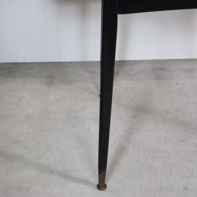 Mid-century British teak dining table leg with slender tapered black finish and brass-capped foot on concrete floor.