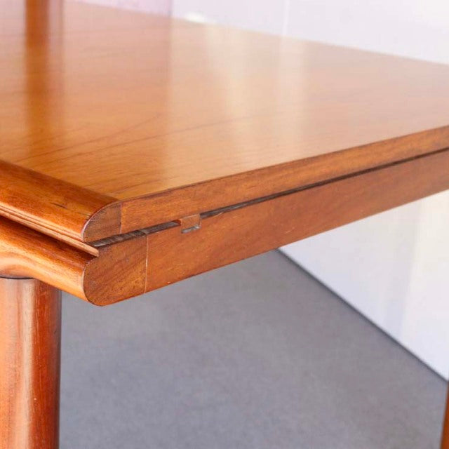 Mid-century British teak dining table corner with smooth wood grain, rounded edge, and sturdy cylindrical leg.