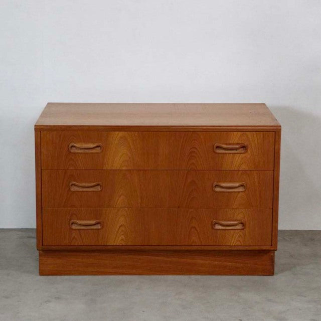 Vintage British mid-century G-plan teak chest with three drawers, curved wood handles, and visible wood grain on clean,.