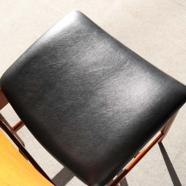 Black leather seat with subtle grain on mid-century British teak dining chair frame, sleek straight legs.