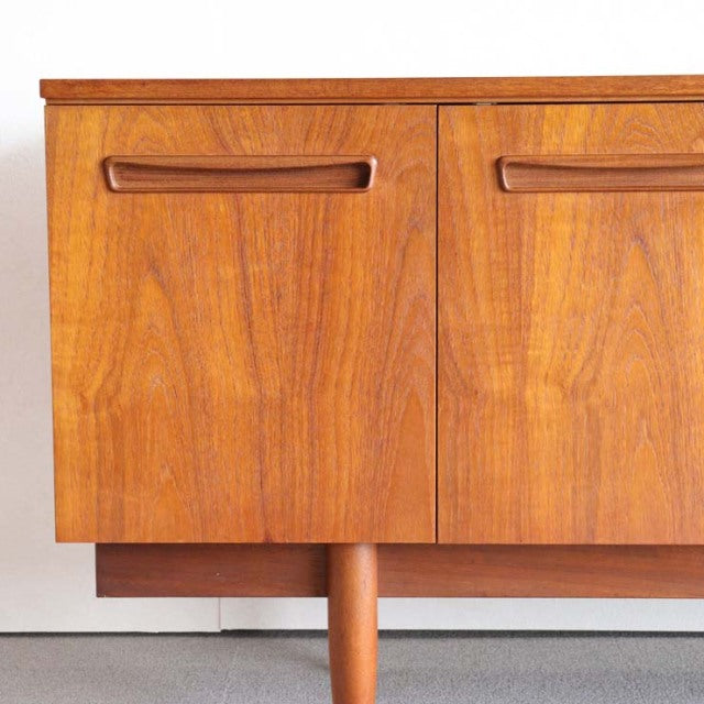 Mid-century vintage teak sideboard with warm wood grain, British-Denmark design, rounded tapered legs, and integrated handles.