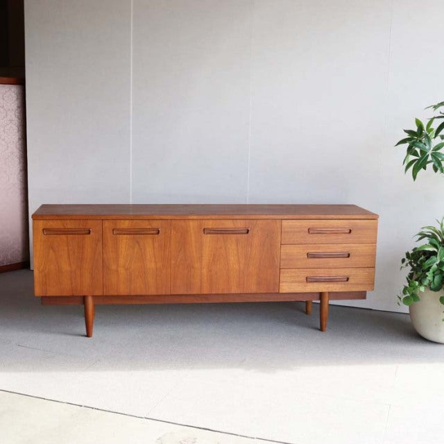 Mid-century vintage teak sideboard with British-Denmark design, featuring wood grain, tapered legs, three drawers, and two.
