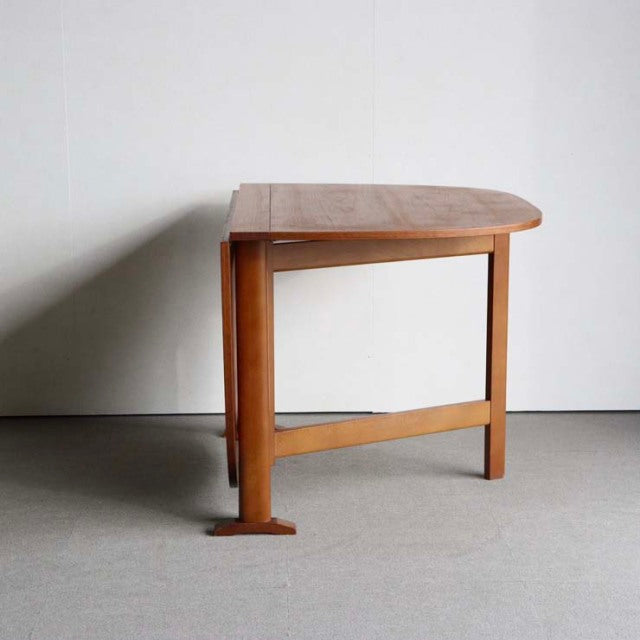 Vintage mid-century British teak folding dining table with rounded corner top and straight legs against plain wall.