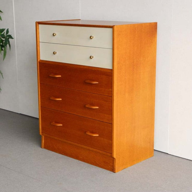 Mid-century British G-Plan teak chest with sunny oak sides and Chinese white top drawers, brass knobs, and sleek wood grain.