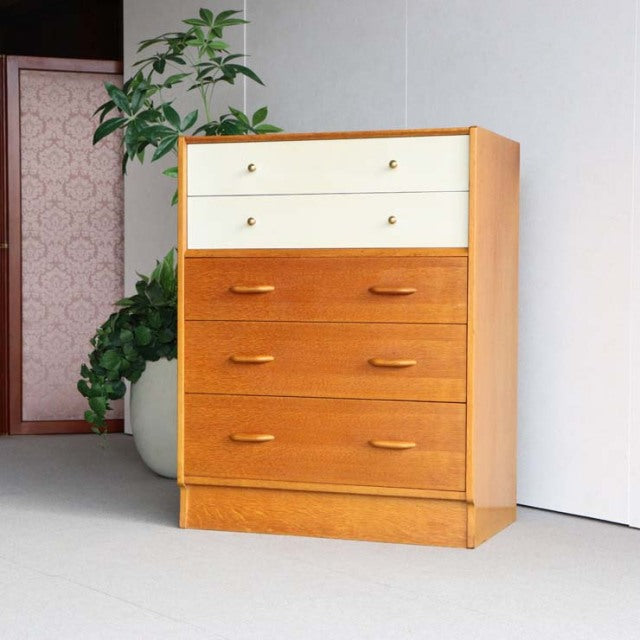Vintage British mid-century teak chest by G-PLAN with warm wood grain, four drawers, and white top drawers with brass knobs.