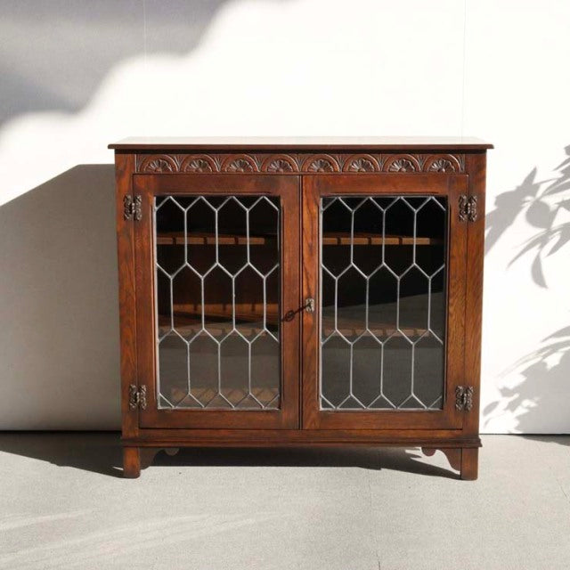 British mid-century vintage teak cabinet by Jaycee with glass-paneled doors and ornate carved detailing atop tapered legs.