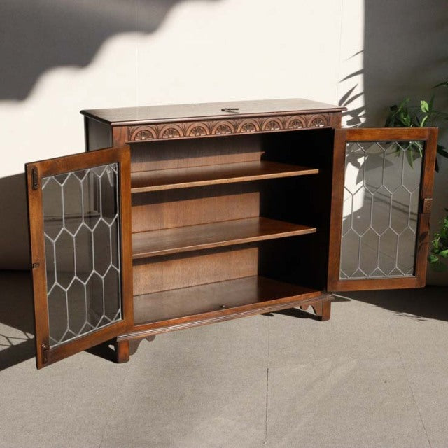 Mid-century British teak cabinet by Jaycee with ornate carved top detail, glass-paneled doors, and tapered legs.