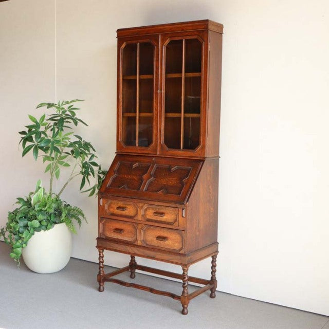 Vintage 1930s teak British antique cabinet with glass doors, intricate wood grain, drop-front desk, and turned legs.