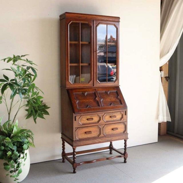 Vintage mid-century British teak cabinet with glass-paneled upper doors, carved wood details, and turned legs.