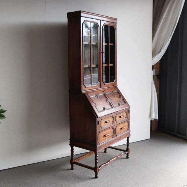 Vintage British teak cabinet from the 1930s featuring glass-paneled upper doors, slanted writing surface, and turned legs.