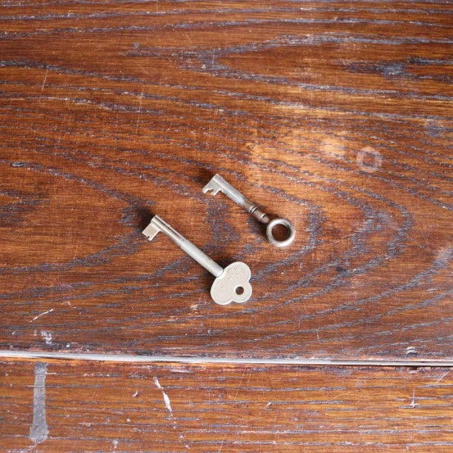 Pair of vintage metal cabinet keys resting on warm teak wood with visible grain, likely from a British mid-century piece.