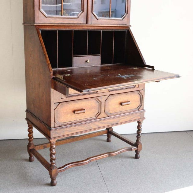 Vintage mid-century British teak cabinet with glass display top, drop-front writing desk, turned legs, and two lower drawers .