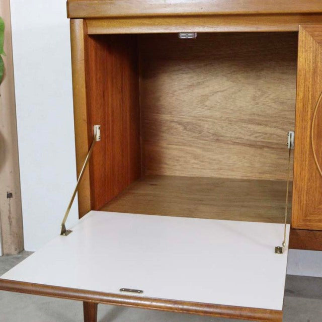 Mid-century British vintage teak sideboard with smooth wood grain, drop-down white front panel, and brass hinge supports.