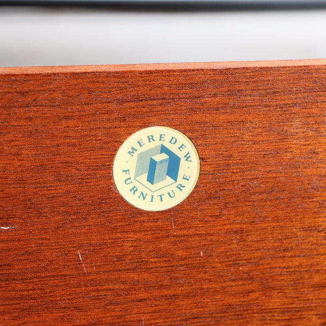 Round cream-colored Meredew Furniture logo sticker on smooth vintage teak wood surface with visible grain.