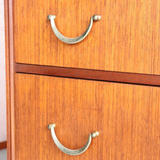 Mid-century teak chest with vintage British design, five drawers, curved brass handles, visible wood grain.