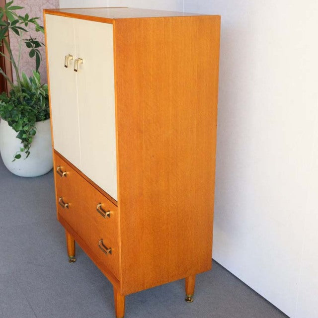Vintage mid-century British G-Plan teak tallboy cabinet with cream doors, wood grain, brass handles, and tapered legs.