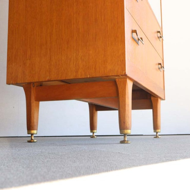 Vintage British mid-century teak cabinet with tapered legs and brass-capped feet, smooth wood grain visible.