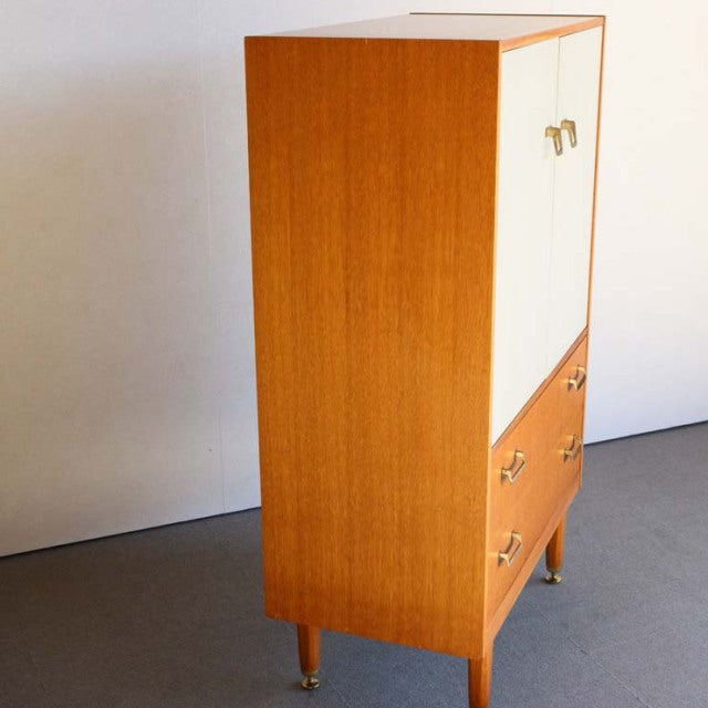 Vintage British mid-century G-Plan teak tallboy cabinet with white doors, brass handles, and tapered legs.