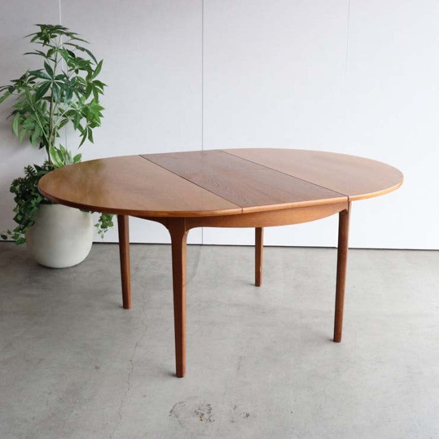 Mid-century British vintage teak dining table with rounded oval top and tapered legs, smooth wood grain finish.