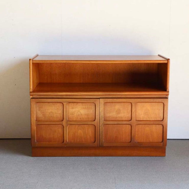Mid-century British teak cabinet by Nathan with cross-design panel doors, rich wood grain, and open top shelf.