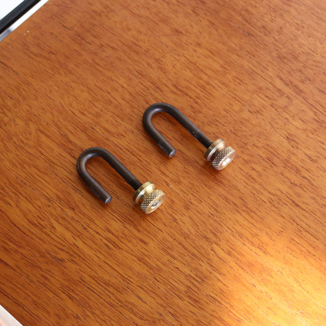Black metal U-shaped hooks with gold-toned screw heads resting on a warm teak wood surface showing fine grain and.