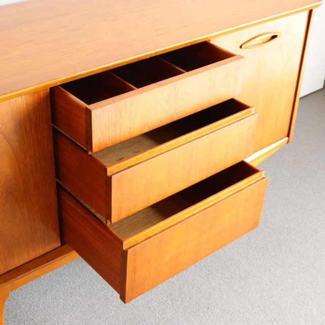 Compact Jentique mid-century British sideboard in teak with smooth wood grain, rounded handles, and tapered legs.