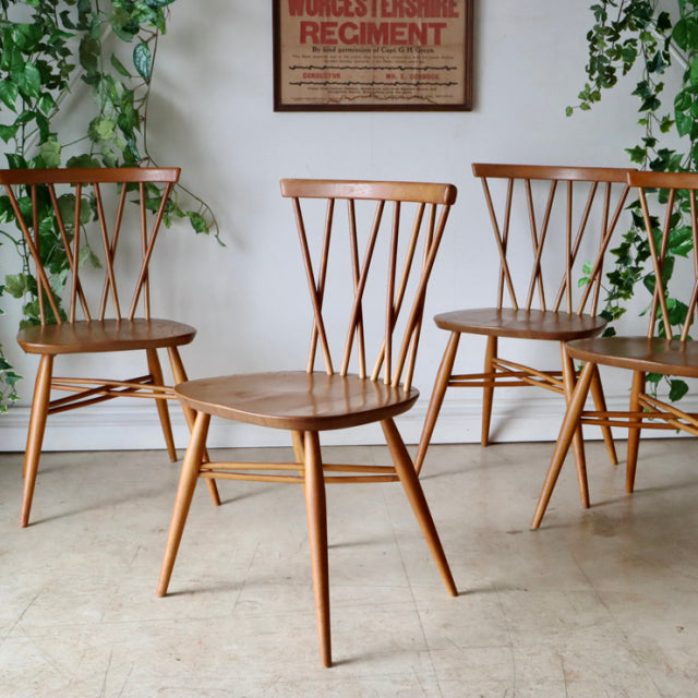 Set of three vintage British mid-century teak X-back dining chairs with spindle backs and tapered legs against a white wall.