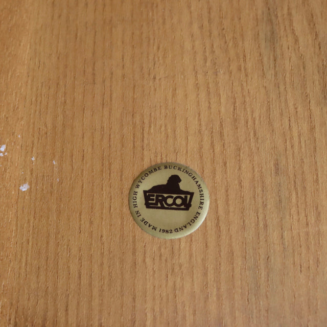 Close-up of round brass Ercol brand badge set into teak wood grain surface, highlighting vintage British mid-century.